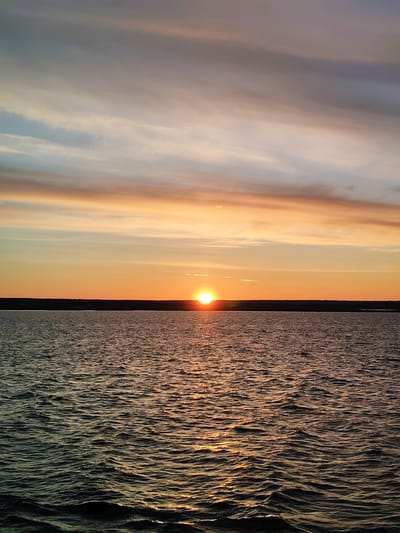 The image depicts a sunset over a body of water. The sun is positioned low on the horizon, casting a bright orange glow across the water's surface. The sky transitions from orange near the horizon to lighter shades of blue and gray higher up, with some bands of clouds present. A dark line of land is visible in the distance, separating the water from the sky. The water itself is rippled, reflecting the sunlight. The location, Vasil'evskoe, Russia, cannot be confirmed from the image alone. No people, other specific objects, or actions are observable.