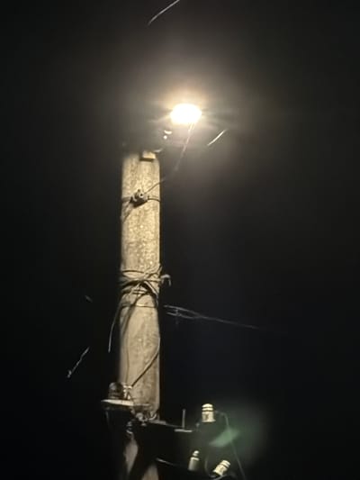 A night-time, low-angle, close-up shot captures a wooden utility pole illuminated by a bright overhead street lamp in Smyadovo, Bulgaria. The pole, weathered and rough, is wrapped with wires and twine, suggesting a rustic or aged infrastructure. The street lamp emits a strong, diffused light, creating a halo effect against the dark, featureless background. At the base of the pole, partially obscured by darkness, are several small, white, cylindrical objects, possibly insulators or other electrical components. The overall impression is one of a solitary, functional object set against the deep black of the night.