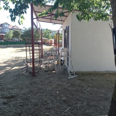 In Çatalca, Turkey, a construction site is depicted. The foreground is dominated by dry, dusty ground covered with straw and debris. Two silver A-frame ladders stand upright against a light-colored wall on the right side of the frame. Behind the ladders, a yellow generator is visible. To the left, a red metal frame structure, possibly a support for a roof or shelter, is partially constructed, with some metal beams lying on the ground. In the background, trees and foliage are present, and further back, a residential building with a red-tiled roof and multiple windows is visible. The overall scene suggests an unfinished construction project in an outdoor, possibly rural or semi-rural, setting.