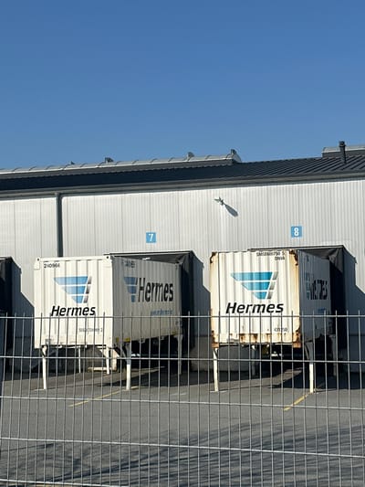 An exterior image depicts a logistics facility loading dock area located in Schloß Holte-Stukenbrock, Germany, under a clear blue sky. The primary structure is a white corrugated metal industrial building with a dark grey roof featuring several vents. Two loading bays are visible, marked with blue numbers "7" and "8" positioned above the respective dock openings, which are equipped with black dock seals.
Two white semi-trailers or shipping containers, prominently branded with the "Hermes" logo (a blue multi-faceted symbol and the word "Hermes" in black), are backed into bays 7 and 8. The trailer at bay 7 displays identifying markings "240966", "4061", "www.myhermes.de", and "SEG|72491 2". The trailer at bay 8 shows markings "SMSE600750 5", "DNVA", and "431250", and exhibits visible rust and deterioration on its lower chassis. Both trailers are supported by their deployed landing gear.
In the immediate foreground, a grey metal mesh fence spans the width of the frame, partially obscuring the asphalt ground, which features faint yellow painted lines. The scene is illuminated by direct sunlight, casting distinct shadows. Notably, a pronounced shadow, resembling a fletched dart or an arrow, is cast onto the white building wall between loading bays 7 and 8. No individuals are present in the image.