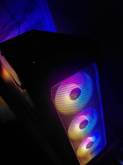 An indoor, low-light photograph captured in Mezdreya, Bulgaria, showcases the top panel of a computer case. The primary subjects are three circular cooling fans, which are brightly illuminated with vibrant RGB lighting, visible through a fine, rectangular-patterned mesh grille. The fans display a dynamic color gradient, transitioning from cool blues and purples on the left to warm pinks, yellows, and oranges on the right, creating concentric rings of light within each fan.
The surrounding environment is predominantly dark, emphasizing the glowing computer components. On the left side of the frame, a distinct blue light spills onto an unseen surface, possibly a floor or wall, where a dark wire is partially visible. This blue illumination adds a cool tone to that area, contrasting with the multi-colored fans. The dark setting suggests the image was taken at night or in a dimly lit room. No human subjects or discernible text are present. The image focuses on the aesthetic and technological appeal of modern computer hardware lighting.