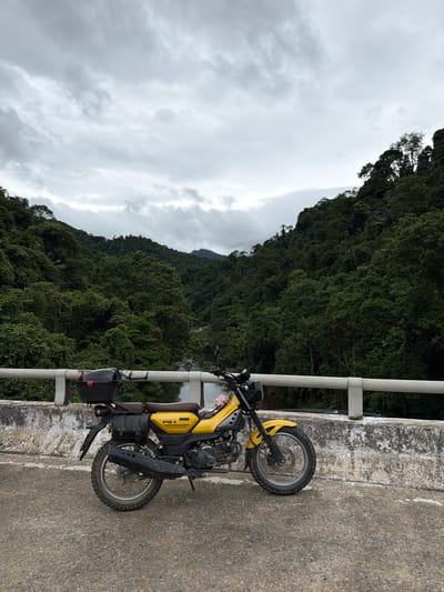 A yellow motorcycle is parked on a bridge in a mountainous, jungle-covered region. The motorcycle, a Honda CT125 Trail 110, is fitted with saddlebags and a top case, suggesting it is equipped for travel. The bridge railing is white and appears to be made of concrete and metal. The background consists of lush green hills and trees, with a cloudy, overcast sky. A river is visible flowing through the valley below the bridge. The scene is likely located in a rural or remote area of Vietnam, possibly in the Kim Ngân Commune, known for its scenic natural landscapes.