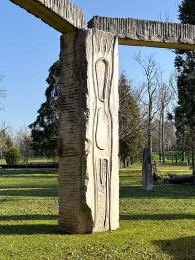 The image displays a large, light-colored stone sculpture located in a park setting within Mealhada, Portugal. The sculpture consists of several monumental stone components, with a prominent vertical pillar in the foreground. This pillar features a smooth, light-beige surface with an abstract, stylized carved relief resembling an elongated, organic form with internal hollowed-out sections. The left side of this pillar exhibits deep, horizontal chiseled striations. A thick, rectangular stone beam with vertical grooves rests horizontally atop the pillar, extending towards the upper left. Another vertical pillar and a horizontal beam with similar textures are partially visible to the right.
The sculpture stands on a well-maintained green lawn, which is evenly lit by sunlight, casting distinct, elongated shadows to the left and rear of the sculpture, indicating the sun is positioned towards the right side of the frame. The background features numerous trees; many are deciduous with bare branches, suggesting a dormant season, while several dark green coniferous trees provide evergreen foliage. In the mid-ground to the right, a solitary, upright grey stone monolith stands, appearing to have a white band or material wrapped around its lower section. Nearby, a large, fallen tree trunk lies horizontally on the grass. In the far left background, a dark park bench is partially visible next to a rounded shrub. The sky above is clear and bright blue. No people are present in the scene.