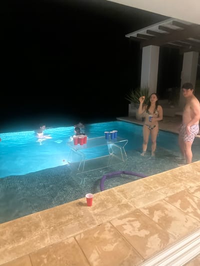 People are playing beer pong in a pool at night. Two players are standing on the side, one with a ball. There are two acrylic tables with beer pong cups arranged on them. One table has red cups; the other has blue. A purple pool noodle lies near one table. A red cup is on the pool deck. Dark and light contrasts exist between the night sky and pool lighting.