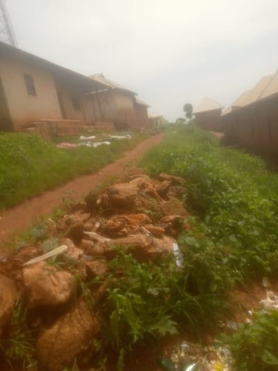 This media file depicts a scene in Mayo Ndaga, Nigeria. The image captures a dirt path winding through an area with several buildings that appear to be under construction or in a state of disrepair. The structures have simple, light-colored walls and metallic or tiled roofs. One building on the left has a visible window. Piles of what appear to be bricks or building materials are stacked near this building, and some white and pinkish items, possibly sheeting or fabric, are scattered on the ground.
The path is bordered by an abundance of green grass and vegetation, which grows thick and somewhat unkempt, with some areas showing small rocks and debris. In the foreground, a collection of larger rocks and dry leaves are visible on the right side of the path, with some scattered trash, including white plastic bags and other refuse, mixed in. Further down the path, more buildings can be seen, including one with a distinct angled roof. A small, sparse tree is visible in the distance, along with another cluster of buildings.
The overall atmosphere is overcast, suggesting a cloudy day, possibly during the rainy season given the lush vegetation and the muddy appearance of the path. The lighting is diffused, and there are no strong shadows, indicating it is likely daytime, but not necessarily bright sunlight. No people are visible in the frame. The scene conveys a sense of rural development or a community in progress, with elements of both construction and untended natural growth. There is no visible text in the image.