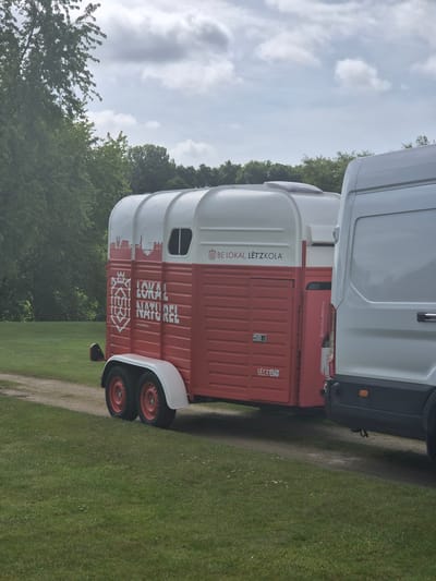 The image shows a white van towing a white and red horse trailer. The trailer is branded with "LOKAL NATUREL" and "BE LOKAL, LËTZKOLA". The setting appears to be outdoors with grass and trees in the background, likely in a rural or semi-rural environment. While I can deduce the "Lëtz" indicates Luxembourg, precise localization within Lenningen requires more specific context.