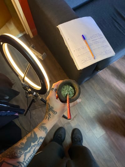 The scene is an indoor setting, likely a home, based on the presence of a grey sofa, wood-patterned flooring, and a ring light. The location is stated as Llavallol, Argentina. A person's arm, heavily tattooed, is holding a traditional South American gourd (mate) filled with yerba mate. A bombilla (metal straw) is inserted into the gourd. The person is wearing a silver ring. To the left of the gourd is a notebook filled with handwritten notes and a pen. A ring light is positioned in the foreground. Black shoes are visible in the background.