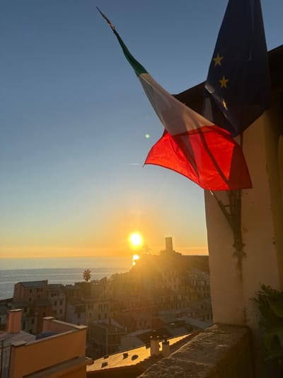 The image presents a panoramic view of Vernazza, Italy, at sunset, observed from an elevated position. In the foreground on the right, two flags are mounted on a light-colored building façade: the Italian national flag (vertical green, white, and red stripes) and the European Union flag (blue field with twelve yellow stars). A rough stone balcony railing is visible below the flags. The background features the town of Vernazza with its multi-story buildings, characterized by terracotta roofs and pastel-colored walls, descending towards the Ligurian Sea. The sun, a bright golden orb, is positioned just above the sea horizon in the left-center, casting strong golden light across the scene and reflecting on the calm water. The Doria Castle tower is discernible on a promontory to the right of the sun, largely silhouetted. Palm trees are visible on the hillsides. The sky is clear, transitioning from light blue overhead to a vibrant golden hue near the horizon. A faint contrail is present in the upper left sky. No individuals are visible.