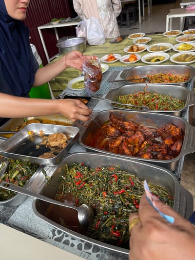 The image depicts an active food service scene at what appears to be a casual eatery or food stall in Kutalhoksukon, Indonesia. A central table, covered with a patterned tablecloth, is laden with a diverse selection of Indonesian dishes presented in both large metal trays and numerous small white plates.

On the left side of the frame, a person wearing a dark blue hijab and a dark shirt is partially visible. Their right arm and hand are prominent as they hold a clear plastic bag filled with a reddish food item, suggesting food is being prepared for takeaway. Nearby, a white plate containing a light green or yellow liquid dish is visible. Another person's hand is visible on the far right, holding a small piece of blue and white paper, possibly indicating a transaction or order. A gold ring is worn on one of their fingers.

The food display includes several large rectangular metal trays holding substantial portions of different meals. One prominent tray contains pieces of chicken coated in a rich, reddish-brown sauce, possibly a chili-based preparation. Adjacent trays contain various stir-fried green vegetables, one featuring water spinach (kangkung) with sliced red chilies, and another appearing to contain green beans or similar elongated vegetables also with chilies. Another tray holds several fried patties, possibly perkedel, amidst some liquid. Metal serving spoons and ladles are present in several of the trays.

Beyond the large trays, an array of small, individual white plates or bowls is arranged across the table, each holding a distinct dish. These include fish in yellow curry sauce, eggs simmered in red chili sauce (balado), and other meat or fish preparations in various sauces.

The background features a reddish-brown corrugated metal wall or partition, indicative of a simple, utilitarian structure. Metal tables and chairs are vaguely visible further back, along with blurred figures of other individuals, suggesting a bustling environment.
