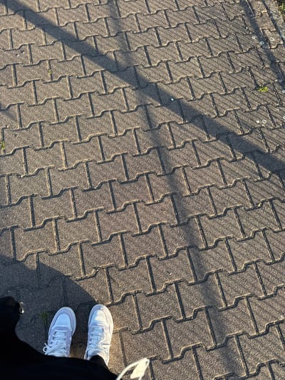 This is a high-angle, full-shot photograph of a person's feet standing on a paved walkway in Bad Säckingen, Germany. The walkway is constructed of interlocking concrete pavers, arranged in a staggered pattern. Sunlight casts long shadows across the pavers, creating a visual texture. The person is wearing white sneakers with purple accents and black pants. The background shows more of the paver walkway and a hint of greenery on the right side, suggesting an outdoor urban environment.