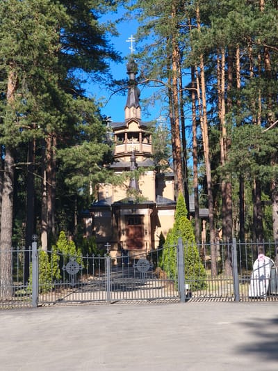 The image shows the wooden Church of St. Sergius of Radonezh in Pesochny, Russia. The church is surrounded by tall pine trees. A gray metal fence with ornate Christian crosses forms the entrance. The pavement in front of the gate leads to the church. A sign is visible to the right of the gate. The church itself is multi-tiered with a dark spire topped with a cross. The sky is clear blue.