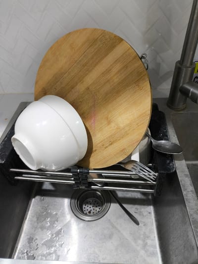 The image shows a kitchen sink with dishes and a cutting board drying. A white ceramic bowl is placed on a metal drying rack. Next to the bowl, a wooden cutting board leans against the sink’s backsplash. A fork and a spoon are also visible on the drying rack. The sink is made of stainless steel and has water droplets and residue on its surface. A drain with a perforated strainer is in the center of the sink. A kitchen faucet is partially visible on the right side of the image. The backsplash is tiled with a pattern that resembles a herringbone design. The scene appears to be in a domestic kitchen, likely after a meal or cleaning. There is no visible text in the image. The lighting suggests it is daytime, but there are no strong indications of the time of day. The overall impression is of everyday domestic life in the Philippines.