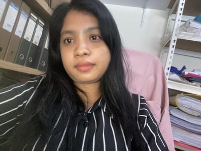 A woman with dark hair is centered in the frame, wearing a black and white striped shirt. The background is cluttered. To the left, a white metal shelving unit is visible, stacked with files and documents. To the right, a row of office binders with labels are arranged on a wooden shelf. The location in the city of Negara, Indonesia cannot be verified.