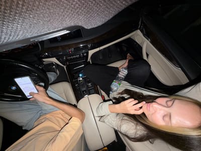 The image depicts the interior of a car at night. Two individuals are visible. In the driver's seat, a person's arm is extended, holding a smartphone which displays what appears to be a data or statistics screen. They are wearing light blue jeans and a light orange shirt. To the right, a woman is seated with her eyes closed, her head resting on her hand, and her hair is partially obscuring her face. She has dyed blonde streaks in her dark hair and is wearing a light-colored long-sleeved shirt. A bottle of water sits on the console between the seats. The car's dashboard and interior are visible, with a dark, possibly leather, trim. The overall lighting suggests it is nighttime, and the location is identified as Sluchainyi, Russia.