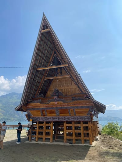 The image shows a traditional wooden stilt house with a prominent triangular roof in Parlondut, Indonesia. The house is ornately decorated with intricate patterns in red, black, and white along the eaves and gable. The roof is made of corrugated metal. The structure is elevated on sturdy wooden posts and appears to be situated on a slight incline overlooking a body of water, likely a lake, with rolling hills and mountains in the background. The sky is a clear blue with a few scattered white clouds. In the foreground, two men are standing and looking at their phones. Another person, partially obscured, is also visible near the house. The ground around the house is a mixture of dirt and dry grass. A wooden fence runs along the edge of the property.