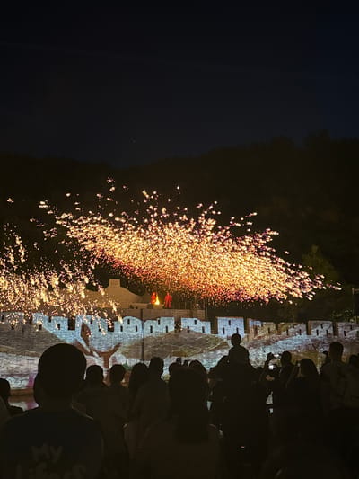 The image shows a nighttime scene in Miyun District, China, featuring a fireworks display near a stone wall. A crowd of people stands facing the wall, many holding up their phones, presumably to capture the event. The fireworks appear as a burst of golden sparks emanating from a source point near the wall. The wall itself shows signs of architectural design, with distinct sections and crenellations. The surrounding environment includes dark areas, likely representing trees or other structures. The dominant action is the fireworks display, with the crowd as observers documenting the event.