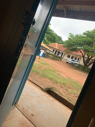 The image captures a view through an open, weathered blue door, looking out onto a courtyard area in Mkar, Nigeria. The foreground is dominated by the interior of the doorway, revealing a blue-painted doorframe and a portion of the door itself, showing signs of age and wear. The floor inside the doorway is made of concrete tiles, cracked in places, with dirt accumulation along the edges.
Beyond the doorway, the scene unfolds into an outdoor setting. A paved path leads into the courtyard, which is covered in a mixture of red earth and patchy green grass. In the middle ground, a single-story building with a terracotta-tiled roof is visible. The building has a cream-colored facade with blue trim around the windows and doors. Several people are seen near the building entrance, some in white uniforms. A silver SUV is parked in front of the building.
The background features lush green trees, suggesting a relatively mild climate. The sky appears overcast, indicating a cloudy day. The overall impression is that of a school or administrative complex, possibly on a rainy or humid day, given the overcast conditions. The scene is framed by the open doorway, giving a sense of looking out from within a structure.