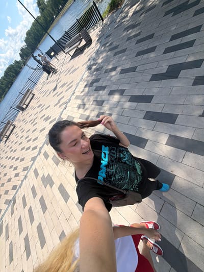 A young woman with dark hair tied in a ponytail is taking a selfie with another person. She is wearing a black t-shirt with a graphic print of the words "DANCE TEENAGE" in blue, and black pants. She is smiling and winking at the camera, holding her ponytail. Next to her, the arm and shoulder of another person are visible, wearing a white top and red shorts, with red sneakers. The scene is set on a paved walkway with alternating light and dark rectangular tiles. In the background, there is a body of water, a black metal fence, and several park benches. Trees line the far bank of the water. The lighting suggests it is a sunny day, possibly late morning or afternoon, casting shadows from the trees and benches onto the walkway. The overall mood appears to be cheerful and relaxed, with the individuals enjoying an outdoor activity. The location context is Белая Холуница, Russia.