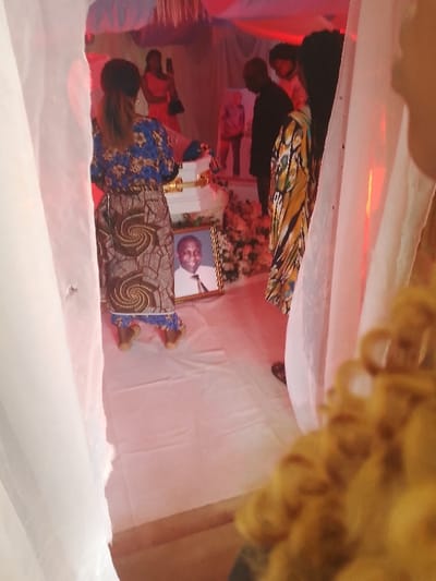 The image depicts an indoor memorial or funeral service occurring in Ufuma, Nigeria. The scene is characterized by a warm, pervasive red ambient lighting. In the foreground, the back of a woman is visible, wearing a blue floral top and a brown patterned skirt with prominent spiral designs. She stands on a white fabric-covered floor. Directly in front of her, positioned on the floor, is a framed portrait of a smiling elderly man, dressed in a white shirt and tie. Behind this portrait, a multi-tiered white structure, resembling a casket or a decorative display stand, is adorned with white and possibly pink floral arrangements.

To the left of the central arrangement, a woman in a pink dress is observed holding a mobile phone, angled as if recording the event. To the right, a man in dark attire stands with his back partially to the viewer, beside another individual clad in a distinctive yellow and black patterned fabric. Additional figures are discernible in the softer focus of the background. The visible area is framed by sheer white drapes or curtains, contributing to a formal and decorated environment.