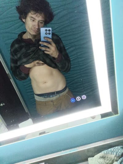 A man takes a mirror selfie. He has curly dark hair and holds a blue smartphone. He is wearing a dark, patterned shirt and brown pants, pulled down to reveal his abdomen. The bathroom has a textured, light blue wall. The illuminated mirror has touch control buttons. A partial view of a dark cabinet is seen on the left.