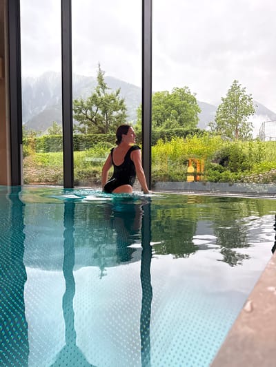 A woman in a black swimsuit is standing in an indoor swimming pool, looking out a large window. The window overlooks a lush green landscape with mountains in the distance. The pool water is clear and reflects the sky and surrounding greenery. The woman is partially submerged in the water, with her hands resting on the edge of the pool. The setting appears to be a luxury spa or hotel in Mayrhofen, Austria, given the mountainous backdrop and modern amenities.