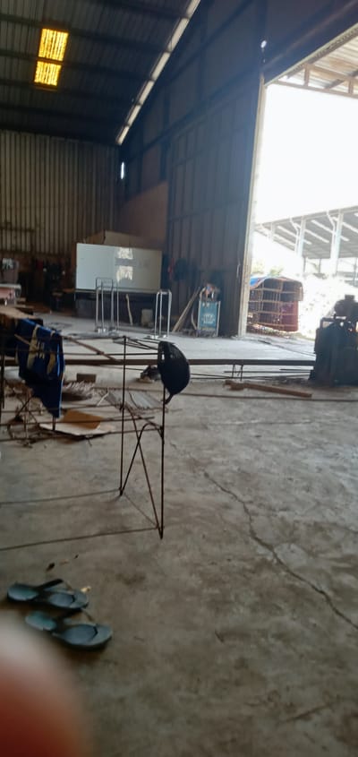 The image depicts the interior of what appears to be a large, open-sided industrial building or warehouse in Sumput, Indonesia. The structure has corrugated metal walls and roof, with some panels allowing natural light in. In the foreground are a pair of rubber flip-flops on a concrete floor. In the mid-ground is a metal stand or frame. The background reveals an open area leading outside to a structure and some foliage. There are also miscellaneous items scattered around, including what seems to be equipment and materials associated with metalwork or construction. No people are visible in the immediate foreground, but some forms exist in the distant background.