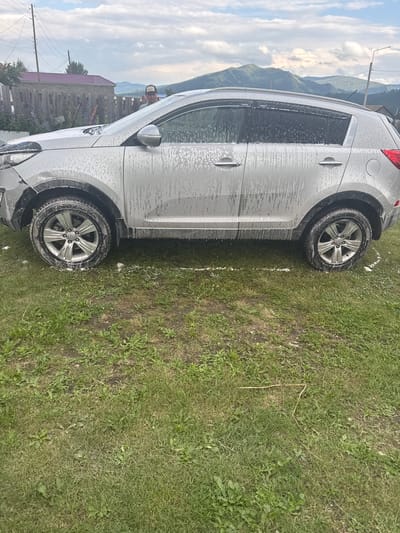 A silver SUV, covered in soap suds, is parked on a grassy area in Yabogan, Russia. The car is facing right. The background shows a wooden fence, a house with a red roof, and a hilly, tree-covered landscape under a cloudy sky. A man wearing a baseball cap is partially visible standing near the front of the car, facing the camera. The scene appears to be set in a rural or semi-rural environment.