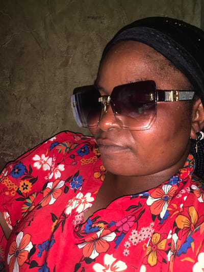 The image shows a close-up, overhead view of a person lying down. The person is wearing a black headscarf and large, dark sunglasses with a metallic accent on the temples. They are also wearing a red dress with a vibrant floral pattern featuring white, orange, and blue flowers. The person's face is partially obscured by the sunglasses and their head is tilted. Their skin appears to be dark, and there is visible sweat on their forehead. The background is a textured, dark surface, possibly concrete or earth. The lighting suggests it may be indoors or during the evening, as there are shadows. The overall impression is one of rest or relaxation. There is no visible text in the image. The provided location context is Boi A, Nigeria.