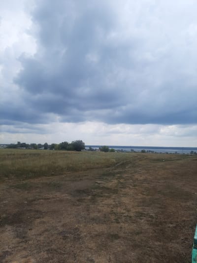 The image depicts a rural landscape with a vast, overcast sky dominating the upper portion. Below the sky, a body of water stretches across the horizon, with a thin line of land separating it from the foreground. Along this distant landmass, a settlement of houses and trees is visible. The middle ground consists of a field of dry, brown grass with a dirt path winding through it. The foreground is also covered in dry grass and patches of bare earth. The scene appears to be a natural, undeveloped area on the outskirts of a settlement.