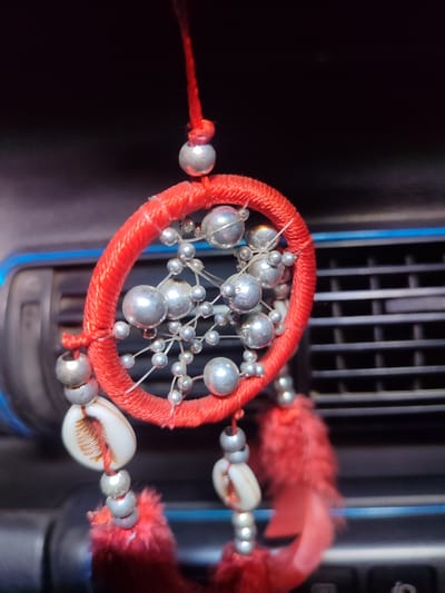 A close-up, macro shot focuses on a small, red-stringed dreamcatcher ornament. The dreamcatcher has a circular red frame adorned with numerous small, silver beads connected by thin wire, resembling a spiderweb. Dangling from the bottom of the dreamcatcher are several strands, each featuring white seashell pendants, silver beads, and fluffy red tassels. The ornament appears to be suspended, possibly from a car's interior, as a dark, textured surface with horizontal slats is visible in the blurred background, consistent with a car air vent. A hint of blue is present on the left edge of the background. The overall lighting is dim, with the focus sharply on the intricate details of the dreamcatcher. The image does not provide any information about the city of Молодёжный, Kazakhstan, nor does it depict any people, actions, or interactions.