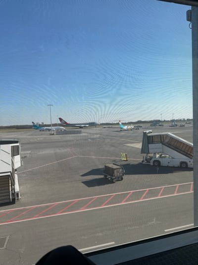 The image shows a view from inside Luxembourg Findel Airport, located at the coordinates provided. The scene is of the tarmac with two planes parked, one a larger white and red plane, and a smaller, turquoise and white plane, positioned at the far end of the tarmac. There is a jet bridge connected to a white airplane at the bottom of the frame.  The scene is captured through a meshed window, with a piece of a person's leg visible in the bottom left-hand corner. There is a red line on the tarmac, and a black baggage cart is parked in the middle of the frame.  The sky is clear blue with a few clouds visible.