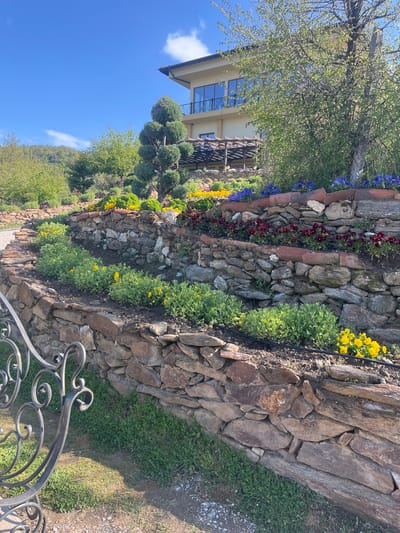 The image depicts a multi-tiered retaining wall constructed of various-sized stones. The wall is situated in a garden setting with a variety of colorful flowers and green plants growing on each level. The flowers include blue, red, and yellow varieties. Behind the wall are several topiary trees. A building with a modern design is visible behind the trees, and a blue sky with some clouds is above. In the foreground, part of a black wrought-iron gate can be seen. The scene has been identified as located in Parvomai, Bulgaria.