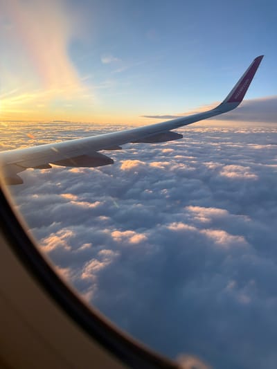 The image depicts an aerial view from an airplane window, looking out over a vast expanse of clouds. The wing of the aircraft, bearing the "wizzair.com" logo, extends into the frame from the right side. The sky is a gradient of blue, transitioning to warmer tones of orange and pink near the horizon, suggesting either sunrise or sunset. The clouds below are illuminated by this light, creating a textured and ethereal landscape. The foreground shows the interior frame of the airplane window. There is no identifiable location in Czerwony Kościół, Poland, as the image is taken from high altitude, showing only clouds and sky.