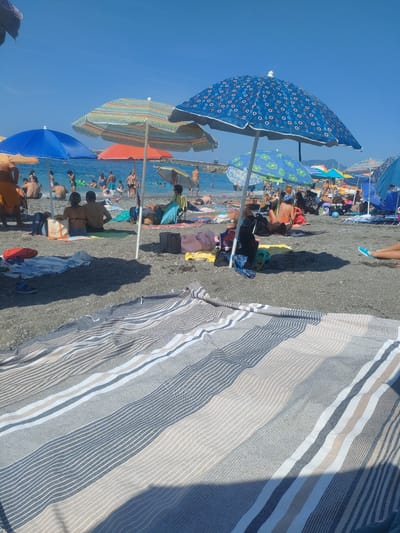 The image depicts a crowded beach on a sunny day in Lavagna, Italy. The foreground is dominated by a large, striped beach blanket laid out on the pebble-strewn shore. In the mid-ground, numerous beach umbrellas provide shade for people lounging and swimming in the clear blue water. The beach is populated with men, women, and children, some sitting on towels, others in the water, and a few walking along the shoreline. Various beach accessories such as bags, a blue flip-flop, and a red object are visible scattered on the sand. In the background, a pier extends into the sea, and hills are visible in the distance under a clear blue sky.