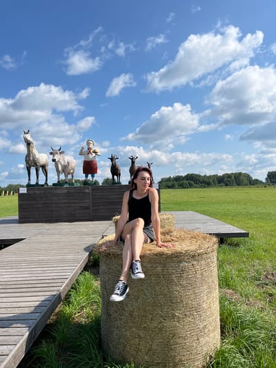 A young woman with dark hair, wearing a black tank top and denim shorts, sits on a hay bale in a grassy field. She is positioned on the right side of the image, with her legs crossed and resting on the bale. Her right hand is on the bale, and her left arm is bent with her hand near her face.

In the background, a wooden walkway leads to a raised platform. On the platform, there are several sculptures: a grey horse, a cow, a chef figure, and two goats. Behind the platform, a green field stretches to a treeline under a blue sky with scattered white clouds. The location is identified as Kurtsevo (Kovriginskii S/S), Russia.