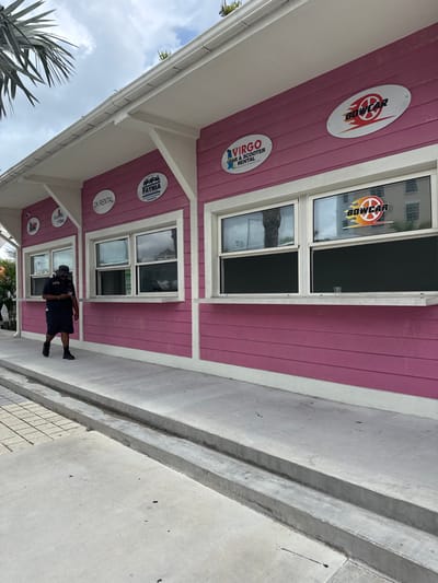 The scene captures a commercial building located in the city of Nassau, Bahamas. The single-story structure is painted pink with white horizontal siding and white trim, featuring a white roof overhang and gutter system. Multiple rectangular windows with white frames and integrated white sills are visible along its facade. Promotional signage on the building includes 'DK RENTAL', 'FATHIA', 'VIRGO CAR & SCOOTER RENTAL', and 'BOWCAR', with an additional 'BOWCAR' decal affixed to a window pane.

On the concrete sidewalk in front of the building, an adult male walks from left to right. He is dressed in a dark short-sleeved shirt, dark shorts, dark shoes, a dark hat, and sunglasses, while looking down at a light-colored mobile phone held in his left hand. A clear plastic cup is positioned on the sill of the rightmost window.

The foreground consists of a concrete sidewalk bordered by a two-tiered concrete channel. Palm foliage is visible at the upper left. Reflections of a multi-story building and vegetation are apparent in the building's windows. The sky overhead is partly cloudy.