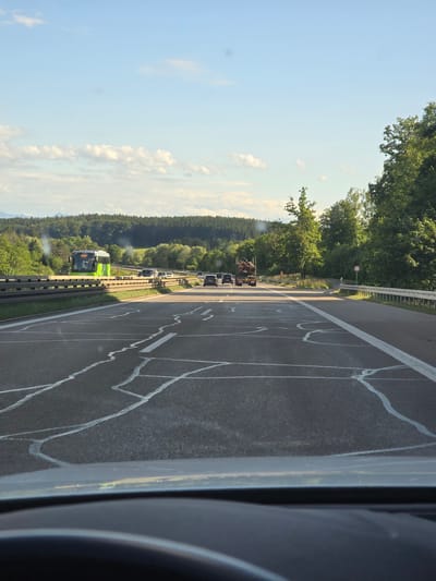 The image depicts a daytime scene on an asphalt highway in Berg, Germany, viewed from the driver's perspective inside a vehicle. The sky is light blue with scattered white clouds, and the sun appears to be low, casting long shadows, indicating late afternoon or early evening.
The highway pavement in the foreground exhibits extensive crack repairs, visible as irregular white lines filling cracks in the dark asphalt. A solid white line delineates the right edge of the immediate lane, and a faded broken white line indicates a lane division further left. The vehicle's dashboard and the top of a steering wheel are visible at the bottom of the frame.
On an elevated bridge or overpass to the left, a green and black Flixbus with "FLIXBUS" text on its side is traveling from left to right. White exhaust fumes or dust are visibly emanating from the rear of the bus. Below the overpass, several dark-colored passenger cars are traveling on a parallel or merging road segment.
Further down the main highway on the right side, a logging truck loaded with timber is visible. Various other passenger cars are present on the highway in the distance. The road is bordered by dense green deciduous and coniferous forests on both sides, with rolling hills or mountains visible in the far background beneath the horizon. A silver metal guardrail runs along the right side of the road, and a circular red and white "no entry" sign is positioned near an off-ramp or restricted access point on the right.