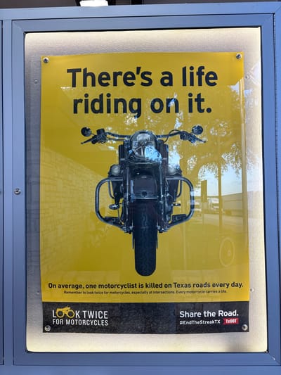 The image shows a close-up of a poster in a public display case in Sonora, United States. The poster has a yellow background and features a large image of a motorcycle in the center. The text on the poster reads: "There's a life riding on it. On average, one motorcyclist is killed on Texas roads every day. Remember to look twice for motorcycles, especially at intersections. Every motorcycle carries a life." Below this text, there is a section with two logos: "LOOK TWICE FOR MOTORCYCLES" and "Share the Road. #EndTheStreakTX TxDOT". The display case has a metal frame and is illuminated from behind. The environment appears to be outdoors, with reflections of trees and vehicles visible on the poster.
