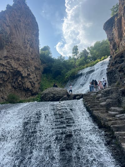 The scene captures the Jermuk Waterfall in Jermuk, Armenia, under a sky with prominent cumulus clouds. The primary subject is a multi-tiered waterfall cascading over dark, stratified rock formations, occupying the central and lower frame. Steep, dark brown, columnar basalt cliffs rise on the left. On the right, rough-hewn stone steps ascend parallel to the waterfall's flow. A group of approximately six to eight individuals, dressed in casual attire, are positioned on the right side of the waterfall, near the stone steps; their actions appear to be observation or traversal. One individual in dark clothing is seated on a large boulder on the left side, while another individual in a white t-shirt and dark shorts stands on smaller rocks nearby. Green trees and dense brush cover the higher ground and crevices between rock faces. Utility power lines are visible stretching horizontally across the upper sky.