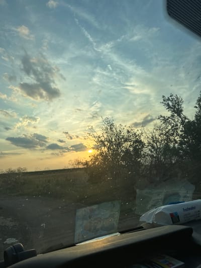 The image is taken from inside a vehicle, looking out through the windshield at a sunset. The sky is filled with a mix of blue and light orange clouds, with the sun peeking through on the right side of the frame. Silhouetted trees line the horizon, obscuring a field and a fence. The dashboard of the vehicle is visible in the foreground, with a package of "Freshmaker" wipes and other assorted items resting on it. The windshield is dusty and shows reflections of the sky and trees. The location is stated to be Mlekarevo, Bulgaria.