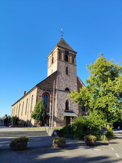 The image shows the exterior of the St. Nikolaus Church in Achern, Germany, on a clear, sunny day. The church is constructed of stone with a tall tower topped by a steeple. Several trees are visible, including a large, leafy tree in the foreground to the right of the church. The church entrance is shaded by a canopy, and there are steps leading up to it. Planters with greenery are placed in the foreground. In the background, a few other buildings and a white scooter can be seen. The sky is a vibrant blue with no clouds.