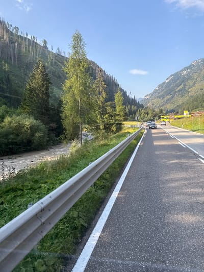 The image depicts a scenic mountain road in Italy, specifically near Mezzavalle, a location within the municipality of Predazzo. The road is asphalt, with a white line marking the edge of the lane. A metal guardrail runs along the left side of the road, separating it from a vegetated embankment and a flowing river.
The river, appearing muddy and turbulent, rushes through a rocky bed, bordered by lush green foliage and trees. On the right side of the road, a sign indicates "MEZZAVALLE". Beyond the road, a hilly landscape unfolds, covered with dense evergreen trees and patches of bare, rocky terrain, suggesting a mountainous region. In the distance, other vehicles are visible on the road, including cars and what appears to be a yellow vehicle, possibly a bus or truck, at a roadside establishment. The sky is clear blue with a few wispy clouds, indicating fair weather. The overall impression is one of a beautiful, natural landscape with a paved road cutting through it.