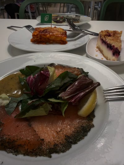 The image depicts a dining scene, likely in a cafeteria or restaurant. In the foreground, a white plate holds a serving of smoked salmon with a dill and herb crust, accompanied by a fresh green salad and a slice of lemon. A creamy yellow dressing is visible on the side. To the left, a fork rests on the plate.
In the middle ground, another white plate contains a piece of lasagna, topped with a green flag featuring a leaf emblem. Next to it are a fork and spoon. A slice of berry cheesecake sits on a smaller white dish to the right, with a crumbly topping and a white filling with visible purple berries.
In the background, out-of-focus green chairs are visible, suggesting a seating area. The overall setting appears to be indoors, on a white tabletop. The presence of multiple dishes and cutlery indicates a meal setting. There are no people clearly visible in the image. The scene does not provide any specific information that would place it in Goyang-si, South Korea.