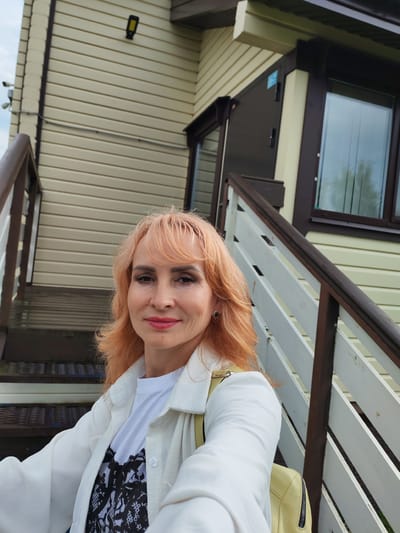 A woman with peachy-colored hair and makeup poses for a selfie on an outdoor staircase. She is wearing a white jacket over a black and white patterned t-shirt and a pale yellow bag is slung over her shoulder. The staircase has wooden steps and white railings, with a darker wooden handrail. Behind the staircase, the wall of a building is visible, clad in horizontal, light-colored siding. A dark brown trim runs around the windows and doors of the building. There is a security camera mounted on the wall to the left of the staircase, and a light fixture above it. The scene appears to be outdoors during the daytime, with cloudy skies visible in the upper left corner of the image. The woman's expression is a slight smile. The setting is likely residential, given the presence of stairs and a house. The location context provided is Kstovo, Russia.