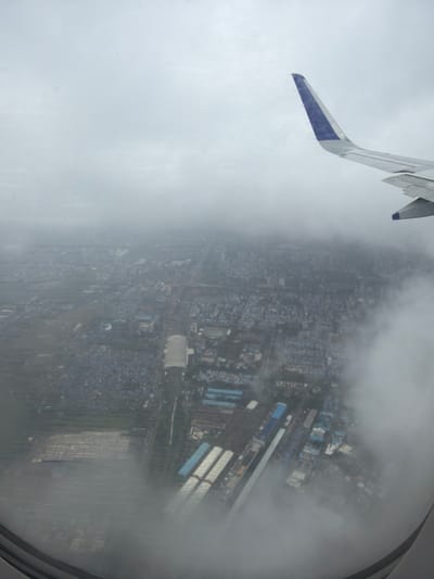 The image is an aerial view from an airplane window, looking down at a dense urban and industrial area, possibly Navi Mumbai, India, under a cloudy sky. An airplane wing, with a blue and white tip, is visible in the upper right corner. The landscape below is characterized by a mix of residential buildings with blue roofs, industrial structures, and some open, possibly agricultural land in the lower left. A large, elongated building with a white roof appears to be a sports stadium or a large industrial complex in the middle left of the frame. Roads and railway lines weave through the city. Clouds partially obscure the view of the ground, creating a hazy and diffused atmosphere. The image depicts a sprawling cityscape under overcast conditions, seen from a high altitude.