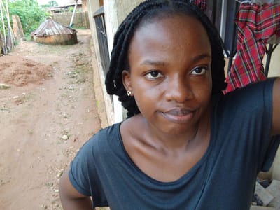 A young Black woman with braided hair and a black t-shirt smiles slightly at the camera. She is wearing a small gold heart-shaped earring in her left ear. She is outdoors, with a dusty path and some greenery in the background. To her right, red and black plaid fabric hangs from a railing. The scene appears to be in a rural or semi-rural area, with what looks like a thatched-roof hut in the distance. The lighting suggests it is daytime, possibly morning or afternoon. The woman's expression is calm and pleasant, and the overall mood of the image is relaxed.