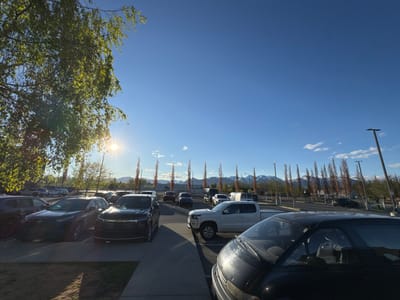 The image depicts a parking lot in Anchorage, United States, on a sunny day. The sky is a clear blue with a few scattered clouds. In the foreground, several cars are parked, including a black SUV, a white pickup truck, and a dark-colored minivan. In the background, there are more parked cars, a row of tall, thin trees, light poles, and a distant view of mountains with snow-capped peaks. The sun is shining brightly through the branches of a tree on the left side of the frame. The overall scene suggests a typical day in a suburban or commercial area of Anchorage.