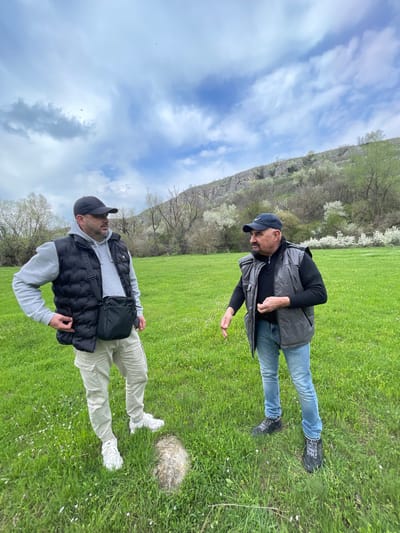 Two men are standing in a grassy field near Barkach, Bulgaria. The first man is wearing a black long-sleeved shirt, blue jeans, a grey vest, a black baseball cap, and black shoes with white laces. The second man is wearing a light grey hooded sweatshirt, khaki pants, a black and grey vest, a black cross-body bag, a black baseball cap, and white sneakers. A rocky, tree-covered hill rises in the background, with a cloudy sky above.