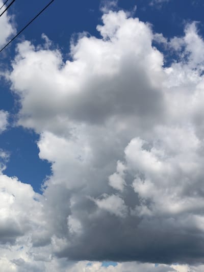 The image shows a sky filled with dramatic clouds. The clouds range from bright white to dark gray, suggesting a mix of sunlight and impending rain. The sky itself is a vibrant blue, visible in patches between the clouds. In the upper left corner, a thin black wire, likely a power line or telephone wire, stretches diagonally across the frame, adding a man-made element to the natural scene. The overall impression is one of a dynamic and somewhat moody sky. The clouds appear to be cumulus or cumulonimbus formations, indicative of unsettled weather. The light suggests it could be daytime, possibly late afternoon given the shadows within the clouds. There is no text visible in the image. The setting appears to be outdoors, with the sky as the primary subject.