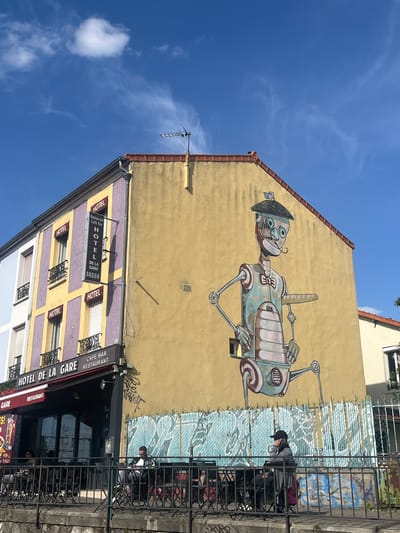 This media file captures a vibrant street scene in Vitry-sur-Seine, France, under a clear, bright blue sky with a few wispy clouds, indicating daytime.
The primary subject is a large, whimsical mural painted on the yellow side of a building. The artwork depicts a stylized, robot-like character with a human-like face, large eyes, and a hat. Its body and limbs appear mechanical, constructed from various metal components, rendered in shades of blue, grey, and brown.
Below this yellow building, another structure features a facade with alternating purple and yellow sections, dotted with windows adorned with black wrought-iron railings. This building houses commercial establishments, prominently featuring the "HOTEL DE LA GARE," which also advertises itself as a "Restaurant Café Bar." Several signs bear these names, with one displaying a phone number: "01 46 82 65 99."
In the foreground, behind black metal railings, people are visible sitting at outdoor tables, suggesting patrons of the "Café Bar Restaurant." One individual, wearing a black cap, is clearly seen seated at a table. Some graffiti is present on the lower part of the yellow wall, behind the outdoor seating area. An antenna is visible on the rooftop of the building with the mural.